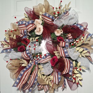 Puede incluir: Una corona patriótica decorada con cinta roja, blanca y azul, arpillera y malla dorada. Presenta una cinta roja, blanca y azul con estrellas, rosas rojas y bayas blancas.