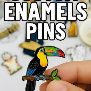 May include: A toucan enamel pin with a yellow chest, rainbow beak, and blue wings, perched on a branch. The pin is held in a hand, with the words "CUSTOM ENAMELS PINS" in white block letters above. Other pins are visible in the background.