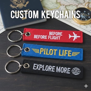Peut inclure: Trois porte-clés personnalisés avec texte brodé et anneaux métalliques. Les porte-clés sont rouges, bleus et noirs. Le porte-clés rouge indique "BEFORE FLIGHT" avec un avion. Le porte-clés bleu indique "PILOT LIFE" avec des ailes. Le porte-clés noir indique "EXPLORE MORE" avec un globe.