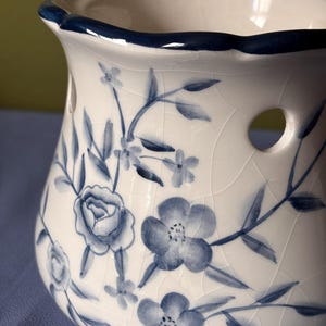 Puede incluir: Un calentador de cera de cerámica color crema con un patrón floral azul y un acabado de esmalte craquelado. El recipiente tiene un borde festoneado con una orilla azul oscuro y dos aberturas circulares en los lados para la ventilación del diseño.
