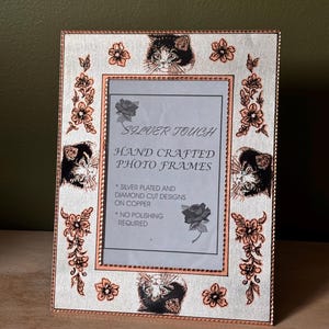 May include: A rectangular silver-plated photo frame with copper-coloured edges and diamond-cut designs. The frame features engraved illustrations of cat faces and floral patterns. The centre insert reads: SILVER TOUCH HAND CRAFTED PHOTO FRAMES, SILVER PLATED AND DIAMOND CUT DESIGNS ON COPPER, NO POLISHING REQUIRED.