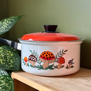 Può includere: Pentola in smalto bianco con coperchio arancione e manico nero. La pentola presenta un fantasioso design a fungo nei toni dell'arancione, marrone e bianco. Un'aggiunta affascinante a qualsiasi cucina.