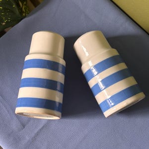 May include: A pair of cylindrical ceramic shakers featuring a glossy off-white base with horizontal blue stripes. Each shaker has a smooth, tapered top section and a wider base, designed for dispensing salt and pepper with a simple, clean aesthetic.