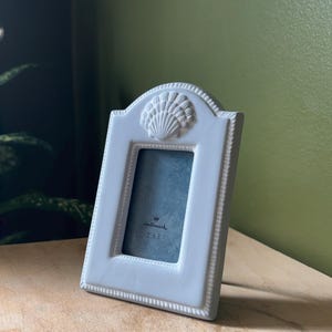 Könnte beinhalten: Ein weißer rechteckiger Bilderrahmen mit abgerundeter Oberkante und einem skulpturalen Muschelmotiv. Der Rahmen hat einen Perlenrand und enthält eine Einlegekarte mit dem Hallmark-Logo und dem Text 5 x 8 cm in der Mitte.