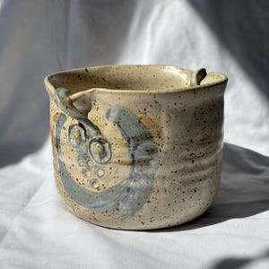 Op de afbeelding: Een beige keramische plantenpot met een blauw en bruin ontwerp. De pot heeft twee handvatten aan de zijkanten.