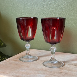 May include: Two matching wine glasses featuring deep red glass bowls with clear vertical cut-out panels. Each glass has a clear, faceted stem and a circular base. The design showcases a contrast between the vibrant red colour and transparent glass.