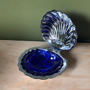 Puede incluir: Un joyero con bisagras en forma de concha marina con un borde exterior de plata metálica y un interior de color azul cobalto profundo. La tapa está levantada, revelando un interior plateado reflectante con un patrón estriado.