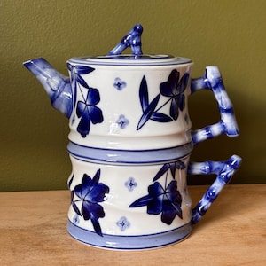 May include: A white ceramic teapot with a stacked bamboo design, featuring blue floral patterns and small star-like accents. The handle and spout are shaped like bamboo stalks, and the lid includes a small bamboo-shaped handle on top.