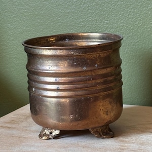 May include: A cylindrical brass planter featuring horizontal ribbed detailing along the upper body and three small, ornate feet at the base. The metal surface displays a warm, aged patina with subtle variations in color and texture throughout.