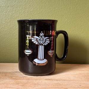 May include: A black ceramic mug featuring a white and gold graphic of a scale of justice centred on the front. Above the scale, the text reads 24.sep.-23.okt. and the word WAAG is partially visible in gold lettering to the right of the scale.