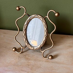 May include: An oval-shaped picture frame with a metallic gold finish. The frame features a decorative border with small leaf-like engravings and is supported by two curved metal stands on each side, each ending in a small spherical finial.