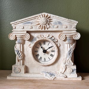 May include: A cream-colored mantel clock featuring a neoclassical architectural design with two ionic columns, a pediment with a sun motif, and draped fabric accents. The clock face has black Roman numerals and the text FIGI printed in the center.