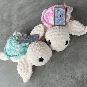 Puede incluir: Dos peluches de tortuga hechos a mano con ganchillo. Una tortuga tiene un caparazón azul claro y blanco, la otra tiene un caparazón rosa y blanco. Ambas tortugas tienen cuerpos, aletas y cabezas color crema con ojos negros. Cada tortuga tiene una etiqueta.