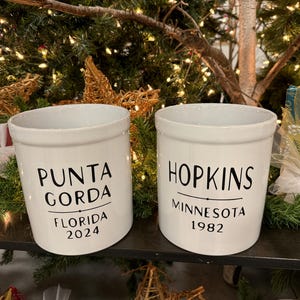 May include: Two white ceramic crocks with black lettering. The crock on the left says "Punta Gorda Florida 2024". The crock on the right says "Hopkins Minnesota 1982".