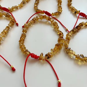 May include: Multiple amber gemstone bracelets with red adjustable cords and gold beads. The bracelets, made of small, irregular stones, are displayed on a white surface. Each bracelet has a sliding knot.