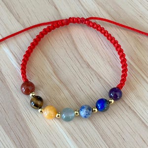 May include: A chakra bracelet with seven gemstone beads in various colours, strung on a red cord with gold-coloured beads. The bracelet is adjustable. The gemstones are in the following colours: red, brown, yellow, green, blue and purple.