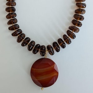 Puede incluir: Collar con cuentas oblongas marrones y detalles dorados. Un gran colgante redondo, rojo-marrón, cuelga en el centro. El collar se asegura con un cierre dorado.