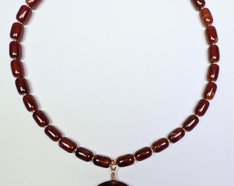 May include: A necklace featuring a strand of dark red, barrel-shaped beads. A large, circular pendant in a matching dark red hue hangs from the center. The necklace is secured with a gold-colored clasp.