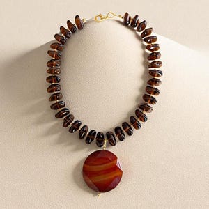 Puede incluir: Un collar de cuentas marrón con un gran colgante redondo rojo y naranja. El collar presenta pequeñas cuentas oblongas marrones ensartadas con un cierre dorado. El colgante es una piedra circular facetada con un gradiente de color cálido.