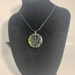 May include: A round, clear pendant necklace with a black cord. The pendant is filled with small, gold-colored beads.
