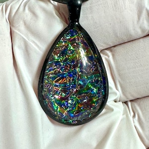 Pode incluir: Um pingente em forma de gota com uma moldura preta e um centro iridescente colorido. O pingente pende de um cordão preto. O centro do pingente tem uma aparência texturizada e multicolorida com tons de azul, verde e dourado.