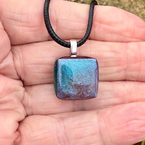 Puede incluir: Un colgante cuadrado con tonos iridiscentes azules y morados cuelga de un cordón negro. El colgante tiene una fianza de color plateado. El collar es una joya.