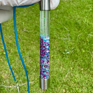 Könnte beinhalten: Eine transparente Glasanhänger-Halskette, gefüllt mit bunten Perlen und Glitzer. Der Anhänger hat silberfarbene Metallkappen und hängt an einer türkisfarbenen Kordel. Die Perlen sind in Lila-, Blau- und Weißtönen gehalten.