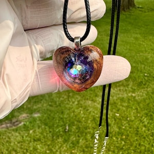 Può includere: Una collana con pendente a forma di cuore e cordino nero. Il pendente presenta un mix di tonalità marroni, dorate e viola, con un centro scintillante e iridescente. La collana è tenuta su uno sfondo erboso verde.
