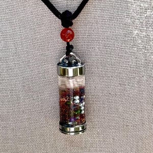 Könnte beinhalten: Eine Halskette mit einem Anhänger aus klarem Glas, gefüllt mit bunten Perlen und kleinen Steinen. Der Anhänger hat silberfarbene Metallkappen und hängt an einer schwarzen Kordel. Eine rote Perle verziert die Kordel über dem Anhänger.