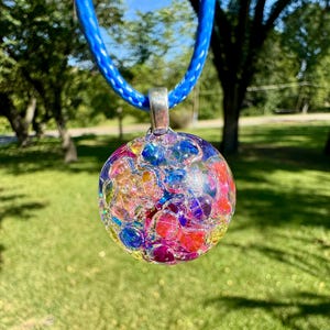 May include: A colorful, round pendant necklace with a blue cord. The pendant is a clear sphere filled with vibrant, multicolored glass beads. The necklace is displayed against a blurred green background.