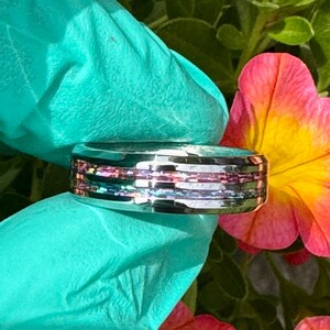 May include: A silver ring with a colorful, layered design. The ring features bands of iridescent and pink hues, creating a unique and eye-catching accessory. The ring is shown against a backdrop of a teal surface and a vibrant pink and yellow flower.