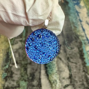 May include: A silver chain necklace with a round blue pendant. The pendant is covered in small, raised blue dots, giving it a textured look.