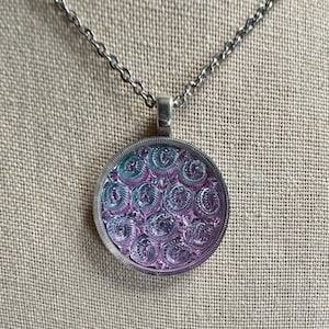 May include: A silver-toned necklace with a circular pendant. The pendant features a pattern of swirling designs in shades of purple and teal, encased in a silver frame. The necklace hangs on a delicate chain, set against a neutral background.