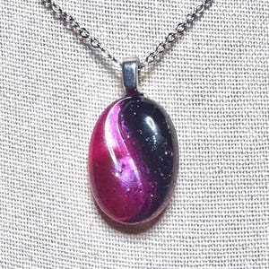 Pode incluir: Um colar com um pingente oval em rosa e preto, com um padrão em espiral. O pingente é suspenso por uma corrente prateada. O pingente tem um acabamento brilhante.