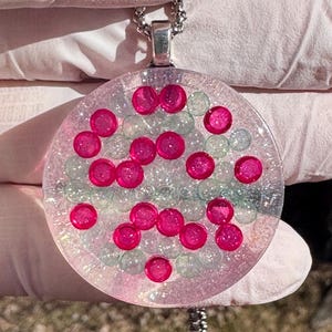 May include: A round pendant necklace with a silver chain. The pendant is clear with pink and light blue beads embedded within. Bright pink, circular beads are scattered throughout the pendant.