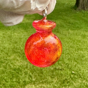 Può includere: Un ciondolo fatto a mano, rosso e oro, con un'ansa color argento. Il ciondolo ha una forma arrotondata con una parte superiore più piccola e svasata. Il ciondolo è tenuto da una mano guantata su uno sfondo verde.