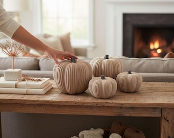 May include: A set of four ribbed, beige-colored decorative pumpkins with dark brown stems. The pumpkins vary in size and are arranged on a wooden surface. Each pumpkin features a rounded, segmented shape with vertical ridges throughout the exterior.