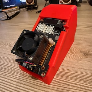 Peut inclure: Un appareil électronique rouge avec un ventilateur noir, des circuits imprimés et un dissipateur thermique argenté. L'appareil porte l'inscription "5VDC G" et "602". L'appareil est posé sur une surface en bois.