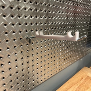 May include: A white metal shelf bracket mounted on a silver pegboard wall.