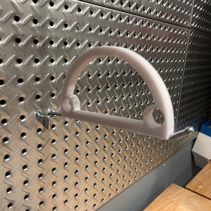 May include: White plastic pegboard hook with a curved design. The hook is mounted on a silver pegboard.