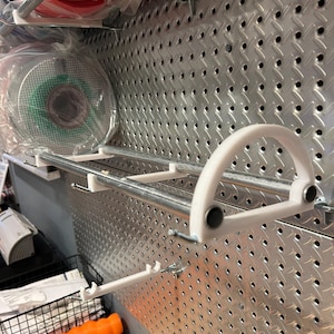 May include: A white plastic wall-mounted storage rack with a silver metal rod. The rack is attached to a silver pegboard wall.