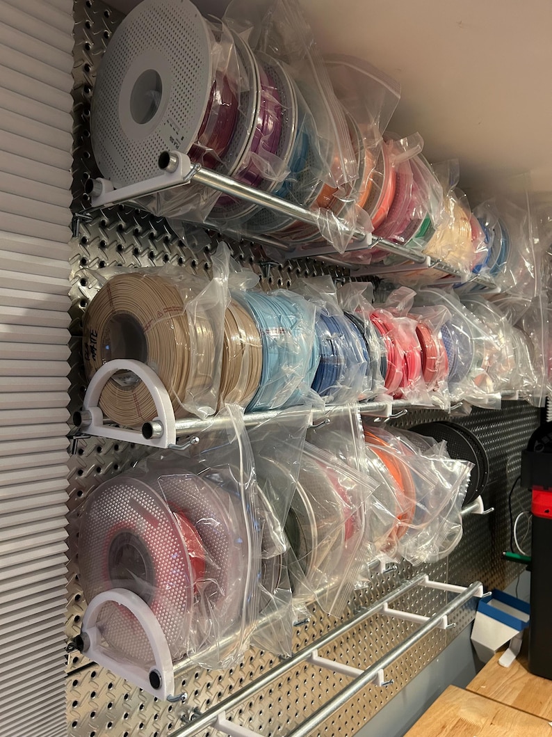 May include: A wall mounted storage system for spools of filament, with multiple rows of spools in clear plastic bags. The spools are various colors, including red, orange, yellow, green, blue, purple, and black.