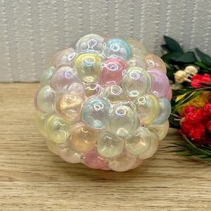 May include: A spherical decorative object made of many iridescent, bubble-like spheres. The colours include pink, yellow, and blue, with a pearlescent sheen. The object is placed on a wooden surface.