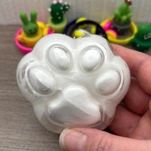 May include: A clear, paw-shaped resin item with a pearlescent, white interior. The paw print has five rounded pads and is held in a hand. In the background are several colorful, small, decorative rings with cartoon figures.