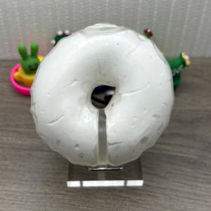 May include: A white, textured donut-shaped object with a hole in the center, mounted on a clear stand. The object is surrounded by small, colorful decorative figurines. The background is a light gray wall and a wooden surface.