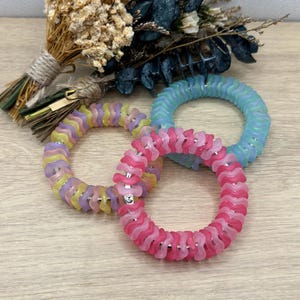 Op de afbeelding: Drie kleurrijke spiraalvormige haarbanden in roze, geel en blauw. Elke band is gemaakt van een flexibel, golvend materiaal en heeft kleine zilveren accenten. Een boeket droogbloemen staat op de achtergrond.