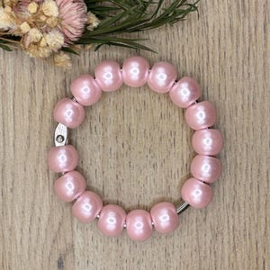 Op de afbeelding: Een roze kralenarmband met een zilveren sluiting. De armband is gemaakt van ronde, lichtroze kralen. De armband wordt getoond op een houten oppervlak met wat bloemen en groen op de achtergrond.