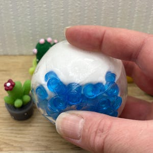 May include: A clear, round stress ball filled with blue gel beads and white foam. The ball is being held by a hand, with miniature cactus figurines in the background.