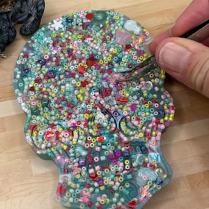 May include: A skull-shaped resin art piece filled with colourful beads, sequins, and small embellishments. The piece is a mix of teal, pink, red, and yellow. A hand with tweezers is working on the piece.