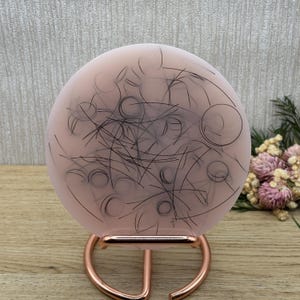 Peut inclure: Un objet décoratif rond rose pâle avec un dessin au trait noir, posé sur un support cuivré. Le motif présente une série de lignes courbes et de cercles, formant un motif abstrait. Le support a un design incurvé et ouvert.